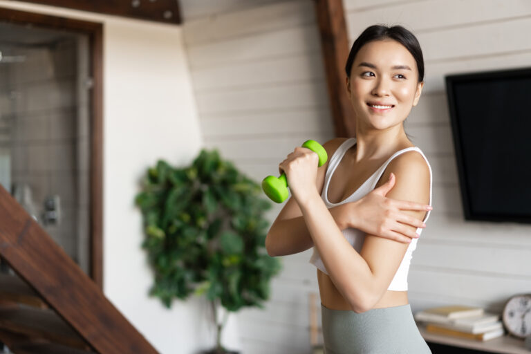 Active and healthy asian girl with fit body doing fitness exercises at home, lifting dumbbells and touching her biceps, workout in living room