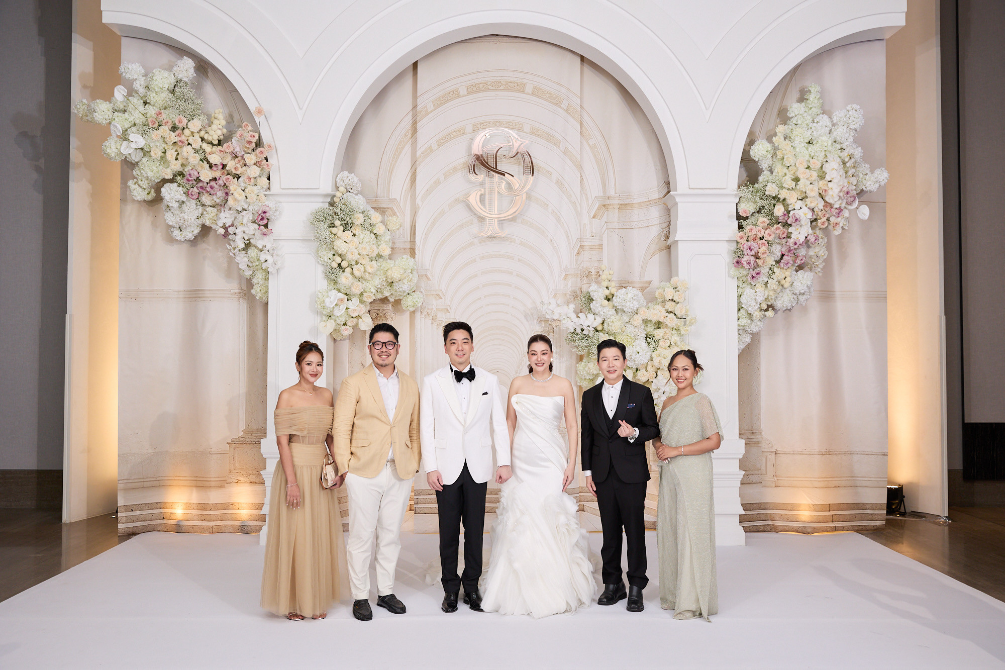 The wedding couple and guests pose for a group photograph in front of an arched backdrop decorated with white flowers.